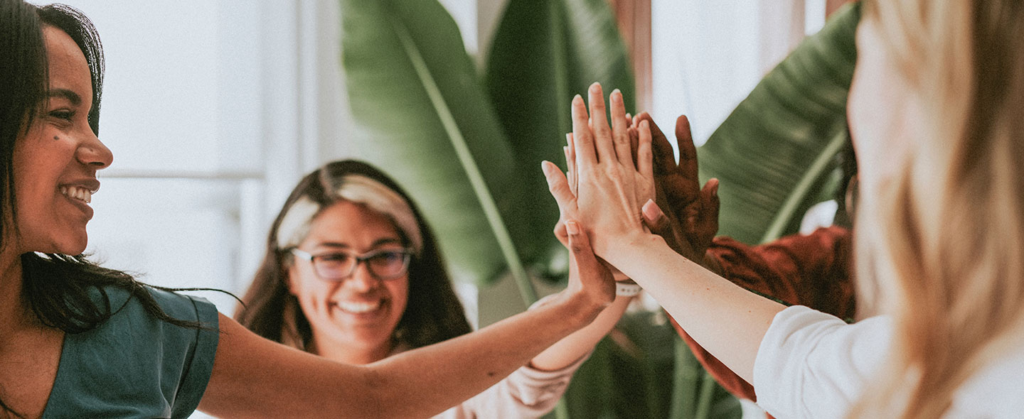 A group of women stand in a circle and reach their hands inwards to give each other a high five.